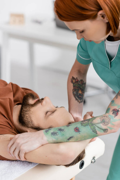 tattooed osteopath massaging injured arm of bearded man in recovery center - Photo, Image
