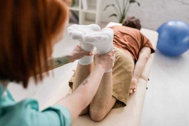 fisioterapista offuscata flettendo le gambe dell'uomo ferito durante la manipolazione di recupero sul tavolo da massaggio in clinica - Foto, immagini