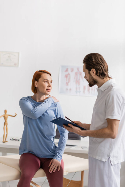 vrouw die pijnlijke schouder aanraakt tijdens overleg met fysiotherapeut die klembord vasthoudt in spreekkamer - Foto, afbeelding