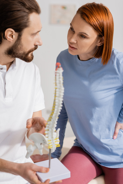 γενειοφόρος οστεοπαθητικός κρατώντας τη σπονδυλική στήλη γυναίκα και μιλώντας με κοκκινομάλλα γυναίκα στο ιατρείο - Φωτογραφία, εικόνα