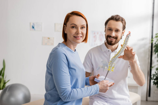cheerful redhead woman and bearded chiropractor looking at camera near spine model in consulting room - Фото, изображение