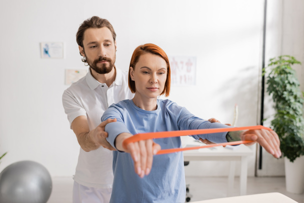 bearded chiropractor assisting redhead woman training arms with resistance band in clinic - Foto, Bild