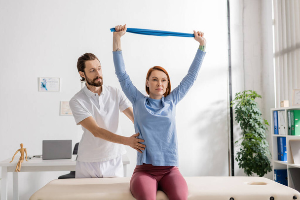 bearded rehabilitation doctor assisting woman sitting on massage table in consulting room and training arms with resistance band - Fotografie, Obrázek
