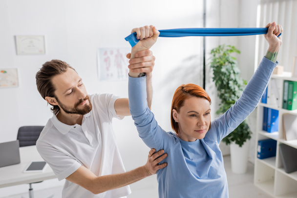 Rehabilitologist assisting redhead woman with resistance band training arms in consulting room - Foto, Imagem