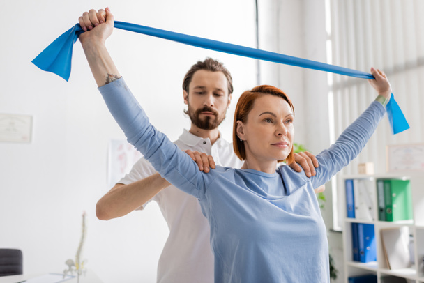 bearded physiotherapist assisting patient working out with resistance band in rehabilitation center - Foto, Imagem