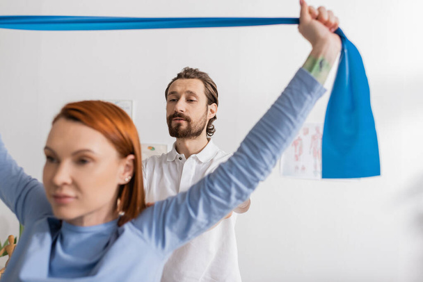 bearded physiotherapist looking at redhead woman exercising with resistance band on blurred foreground in recovery center - Fotografie, Obrázek