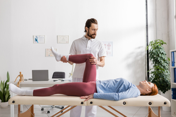 Rehabilitólogo flexionando dolorosa pierna de mujer acostada sobre mesa de masaje en sala de consulta - Foto, imagen