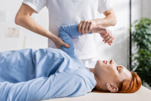 chiropractor flexing injured arm of redhead woman in rehabilitation clinic - Фото, зображення