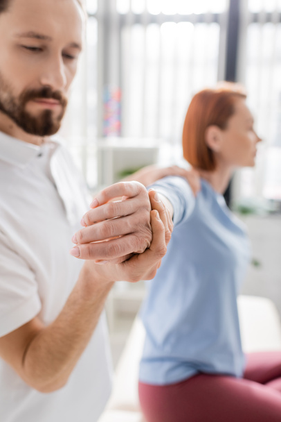 bearded physiotherapist stretching painful arm of blurred woman during diagnostics in consulting room - Φωτογραφία, εικόνα