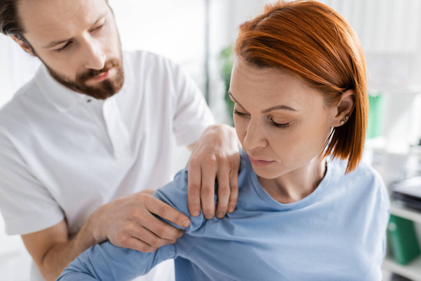 fysiotherapeut raakt gewonde schouder van roodharige vrouw tijdens diagnostiek in spreekkamer - Foto, afbeelding