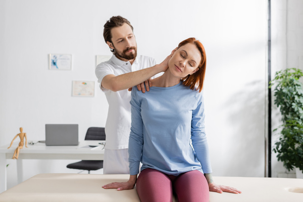 fisioterapeuta barbudo examinando el doloroso cuello de la mujer sentada en la mesa de masaje en la sala de consulta - Foto, Imagen