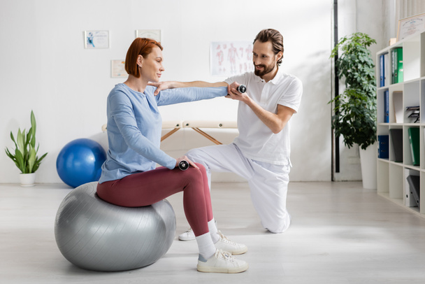 fisioterapeuta sonriente ayudando a la pelirroja sentada en la pelota de fitness y haciendo ejercicio con pesas en la clínica de rehabilitación - Foto, imagen