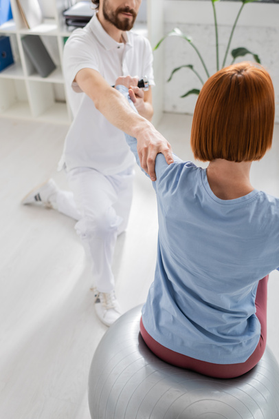rehabilitant podtrzymujący ramię rudej kobiety siedzącej na piłce fitness i ćwiczącej z hantlami w gabinecie - Zdjęcie, obraz