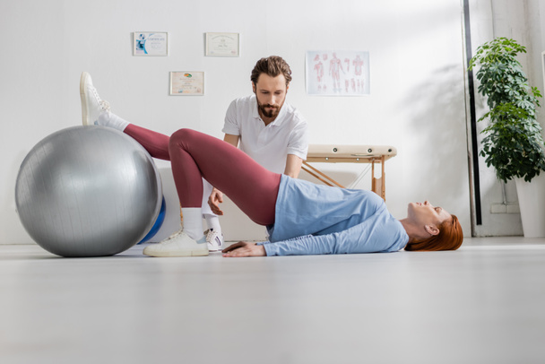 nő fekszik a padlón, és edzés fitball közel szakállas fizioterapeuta rehabilitációs központ - Fotó, kép