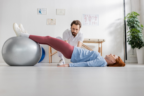 zrzka žena trénink s fitness míčem při ležení v blízkosti fyzioterapeuta v rehabilitačním centru - Fotografie, Obrázek