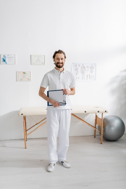 full length of carefree physiotherapist holding clipboard with paper while standing in consulting room - Foto, Bild