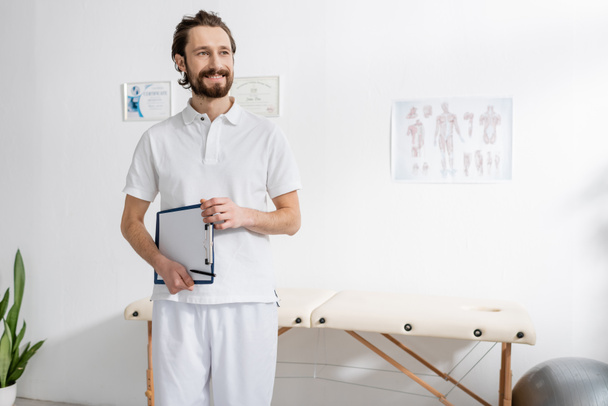 happy manual therapist holding clipboard with blank paper and looking away in rehabilitation center - Fotoğraf, Görsel