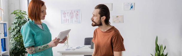tattooed physiotherapist with digital tablet talking to bearded man in consulting room, banner - Photo, Image