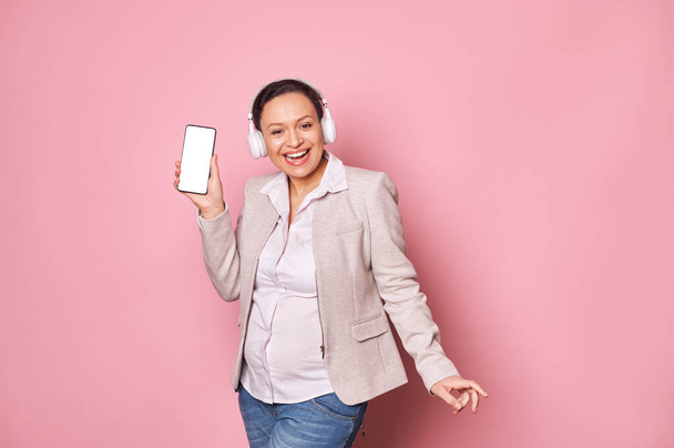 Onnellinen hymyilevä raskaana oleva nainen kuuntelee musiikkia kuulokkeilla, tanssii vaaleanpunaisella taustalla, pitäen älypuhelinta valkoisella tyhjällä digitaalisella näytöllä, jossa on kopiotilaa mobiilisovellukselle. Ihmisiä. Teknologia - Valokuva, kuva
