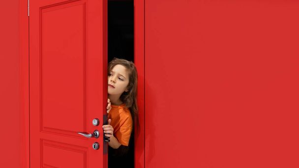Portrait of young little girl, kid peeking out red door and attentively looking, overhearing. Playful and curious mood. Concept of childhood, emotions, facial expression, lifestyle - Foto, Imagem