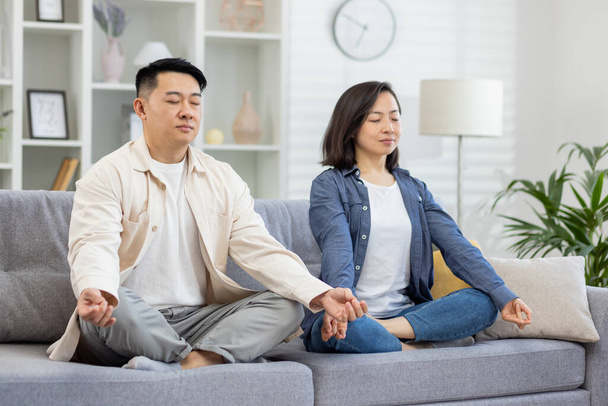 Happy asian family at home on sofa meditating, couple man and woman sitting in lotus position in living room with closed eyes resting relaxing together. - Photo, Image