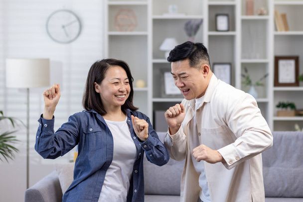 Happy Asian couple man and woman dancing together at home in daytime in living room, happy young family celebrating happy day and wedding anniversary. - Fotografie, Obrázek