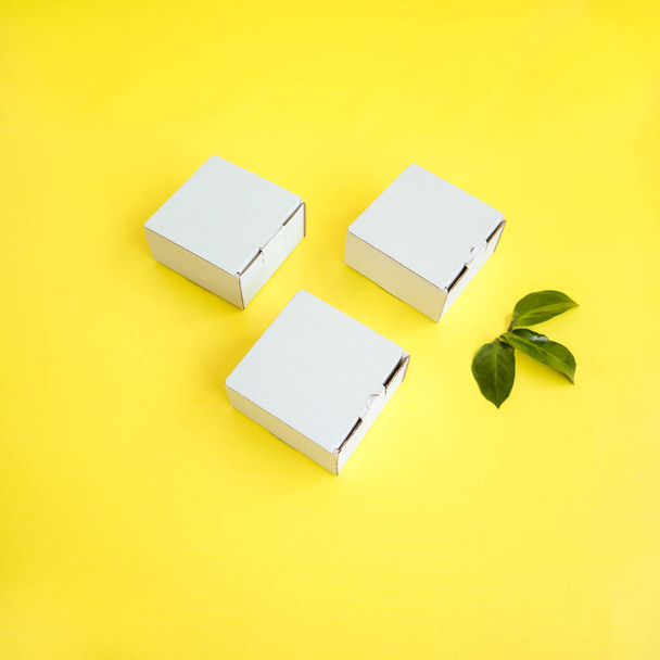 White craft boxes on a yellow background with a decor of green leaves. Top view. Place for text and logo. The concept of a single style of the company, branding, packaging, gifts, holiday, eco style. High quality photo - Zdjęcie, obraz