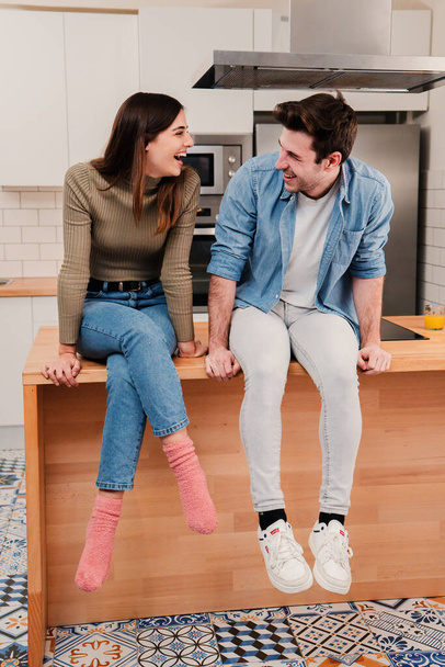 Vertical view of happy young caucasian couple laughing, looking at each other, sitting at kitchen countertop at home. Cherful husband and wife laughing and talkin together resting on their apartment - 写真・画像