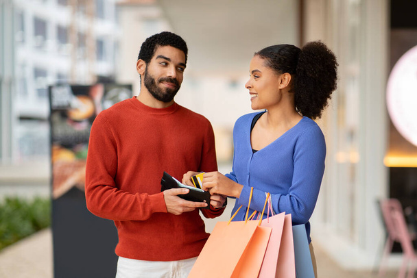 Семейный бюджет. Пара шоппинга вместе, муж дает кредитную карту от кошелька до улыбающейся жены, держащей бумажные сумки в современном торговом центре. Концепция шопаголизма - Фото, изображение