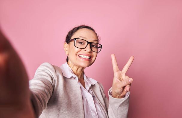 Передний снимок темноволосой счастливой женщины средних лет, мило улыбающейся, показывающей знак мира, делая автопортрет на смартфоне в протянутых руках, на изолированном розовом фоне - Фото, изображение