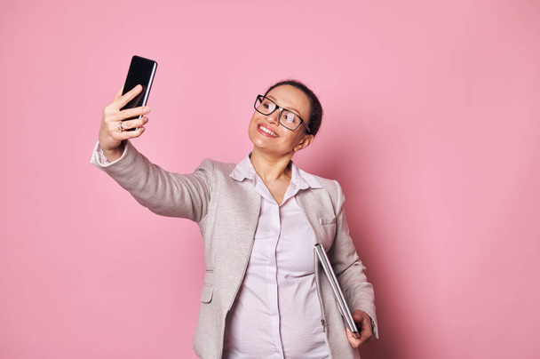 Привлекательная беременная женщина средних лет, бизнесмен в повседневной одежде и модных очках, носит ноутбук и делает селфи на своем смартфоне на изолированном розовом фоне. Рекламное пространство - Фото, изображение