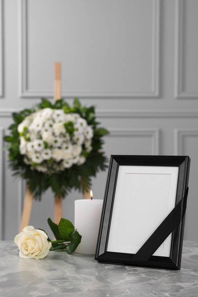 Photo frame with black ribbon, rose, burning candle on light grey table and wreath of flowers near wall indoors. Funeral attributes - Фото, изображение