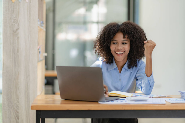 Afroamerykańska bizneswoman w garniturze czyta dobre wieści o młodym mądrym księgowym siedzącym w kolegi patrząc na laptopa Podniosła pięść, wiwatując na sukces swojego projektu. - Zdjęcie, obraz