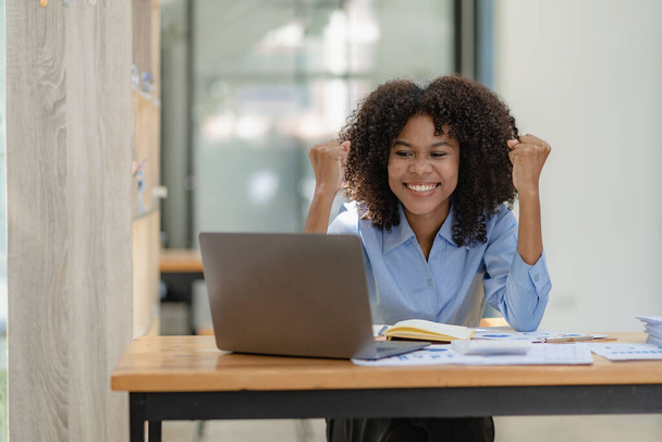 Afroamerikanische Geschäftsfrau im Anzug liest gute Nachrichten über eine junge kluge Buchhalterin, die in einem Kollegen sitzt, während sie auf ihren Laptop blickt. Sie hob die Faust und jubelte über den Erfolg ihres Projekts. - Foto, Bild