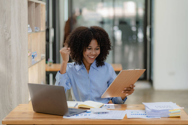 Афроамериканская деловая женщина в костюме читает хорошие новости о молодом умном бухгалтере, сидящем в коллеге, глядя на ноутбук, она подняла кулак, приветствуя успех своего проекта. - Фото, изображение