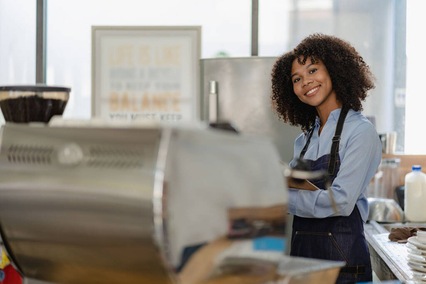 Barista, kahve dükkanı açılışı, Afrikalı Amerikalı bayan girişimci tezgahın önünde duruyor. Önlüklü genç bir bayan garson mağazada müşterilere servis yapıyor. - Fotoğraf, Görsel