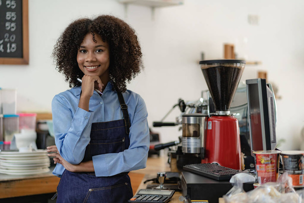Barista, coffee shop startup, African American female entrepreneur standing in front of the counter. A young female waitress in an apron serving customers in a store - 写真・画像