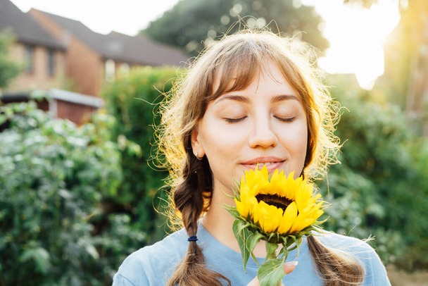 Close up portrait of tender young woman with closed eyes in blue t shirt holding fresh yellow sunflower and enjoying the moment in sunset light. Summertime. Selective focus - Фото, изображение