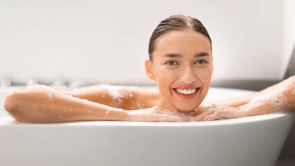 Gorgeous Woman Taking Bath Posing In Bathtub Smiling To Camera In Modern Bathroom At Home. Lady Bathing Caring For Body Covered With Soap Sud. Beauty Routine Concept. Panorama - Foto, imagen