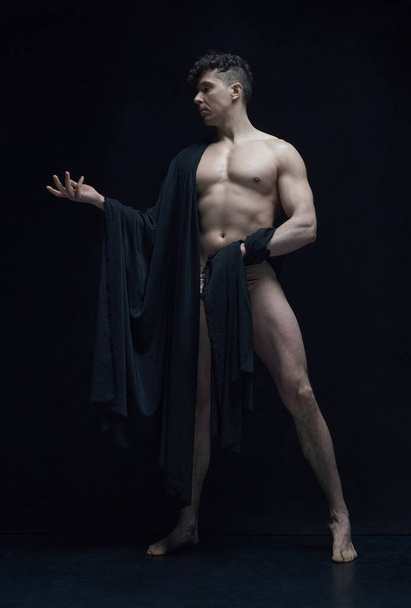 Full-length portrait of muscular, shirtless, handsome man posing in dark fabric against black studio background. Statue art. Concept of male body aesthetics, mens beauty, inspiration, art - Фото, изображение
