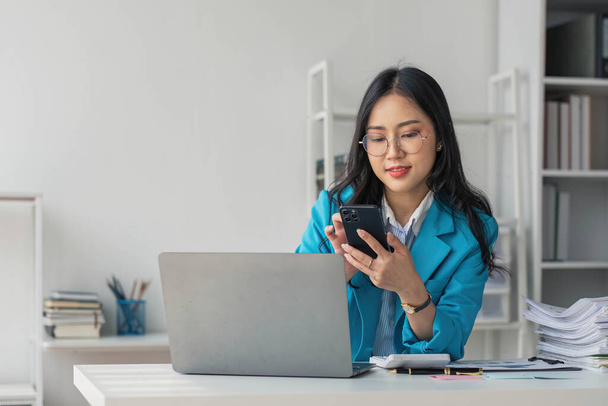 Азійська жінка-підприємець зайнята своєю роботою в офісі. Молода азіатка розмовляє по смартфону чи мобільному телефону під час роботи на комп "ютері.... - Фото, зображення