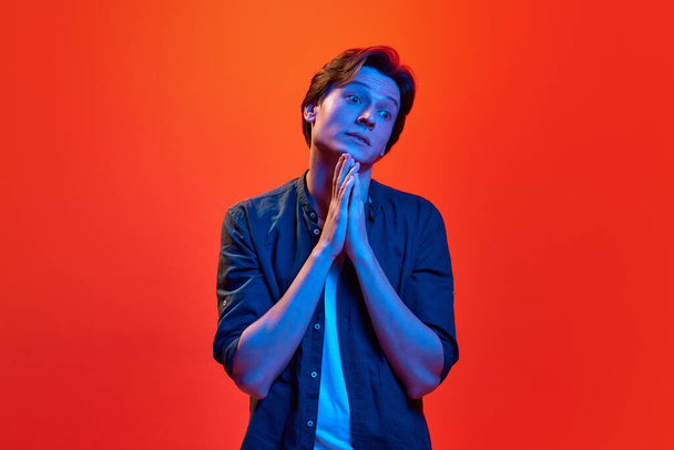 Repentance, plead, implore. Studio shot of young guy, student looking with compassionate eyes over red-orange background in neon light. Concept of beauty, youth, human emotions, mood, ad - Foto, imagen