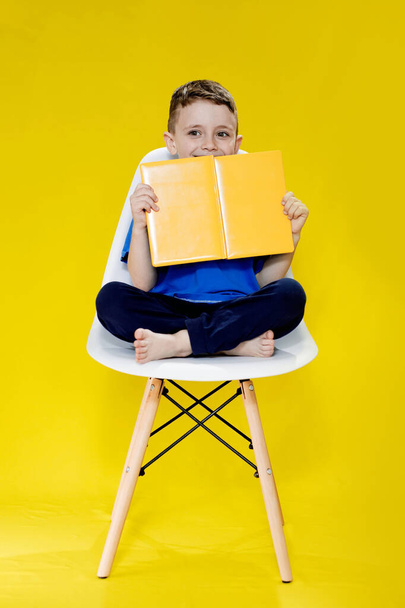 Little cheerful blond green-eyed boy 5-6 years old in a stylish blue T-shirt holding book and reading on yellow wall background, children's studio portrait. - Foto, Imagem