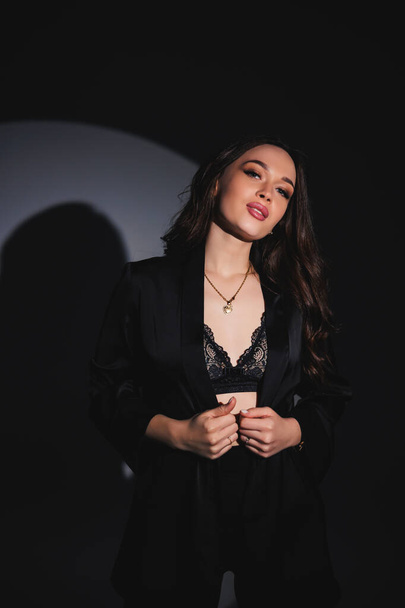 A young beautiful woman in a black suit stands with her hands behind her head on a gray background. Attractive woman in underwear and a jacket - Foto, imagen