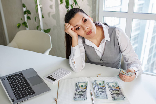 Sad business woman sits at work desk in front of laptop and counts cash. Concept of low profits, low salary and financial problems. - Photo, Image