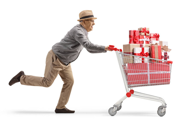 Full length profile shot of an elderly gentleman running with a shopping cart full of presents isolated on white background - Photo, image