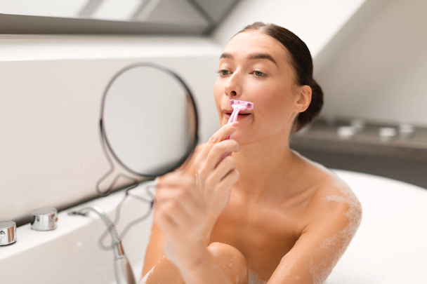 Facial Depilation. Young Lady Removing Face Hair Shaving Moustache Above Lips With Safety Razor, Looking At Round Mirror Taking Bath Sitting In Bathtub In Bathroom At Home. Selective Focus - Photo, Image