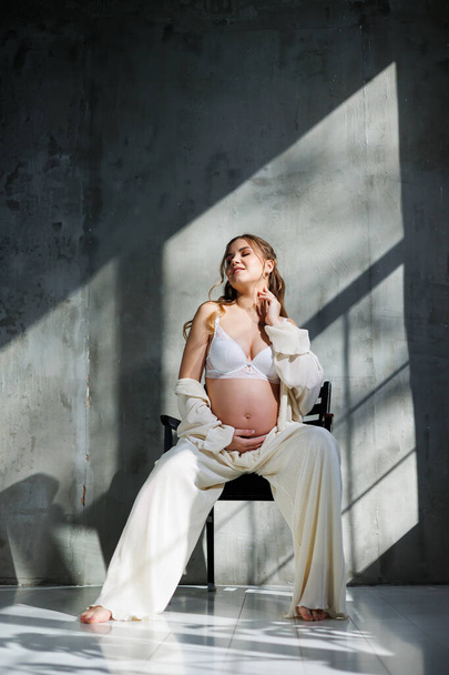 A pregnant pretty woman in a white suit and bra is sitting on a gray background. Pregnant cute woman stroking her belly and smiling. Happy period of pregnancy - Фото, изображение