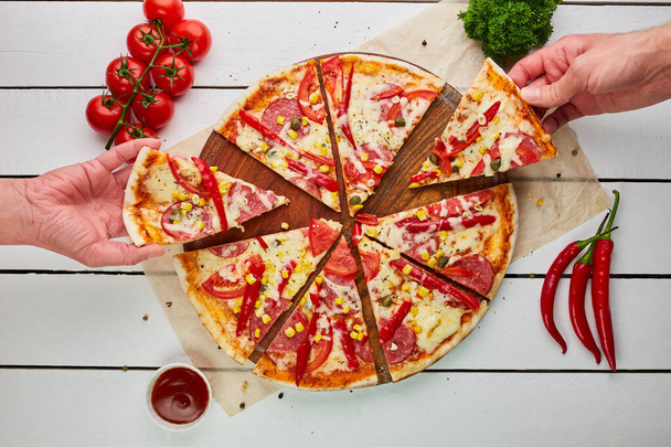 Die menschliche Hand nimmt frisch gebackene leckere Pfefferoni-Pizza mit Salami, Mozzarella-Käse, Mais und Paprika, serviert auf hölzernem Hintergrund mit Tomaten, Soße und Kräutern. Essenslieferkonzept. Speisekarte - Foto, Bild