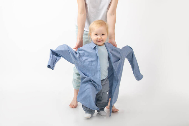 Portrait of amazing family posing on white background. Unrecognizable young woman mother holding hands of wonderful little blond girl baby daughter wear oversized denim shirt. Childhood, growing up. - Photo, Image